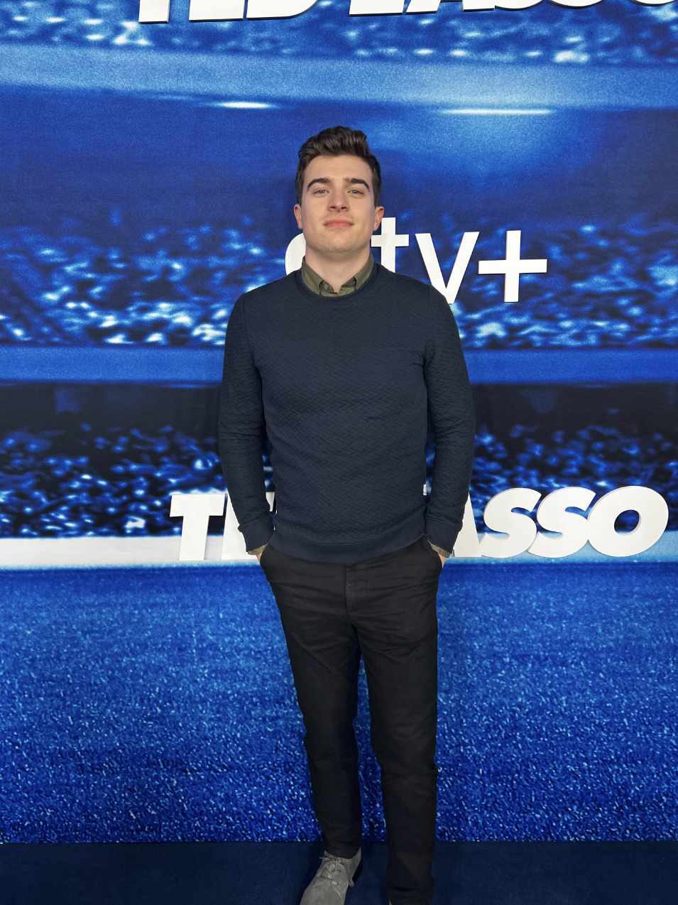 Connor Ripple standing in smiling in black slacks and a blue sweater at the Apple TV+ "Ted Lasso" Season 3 Premiere